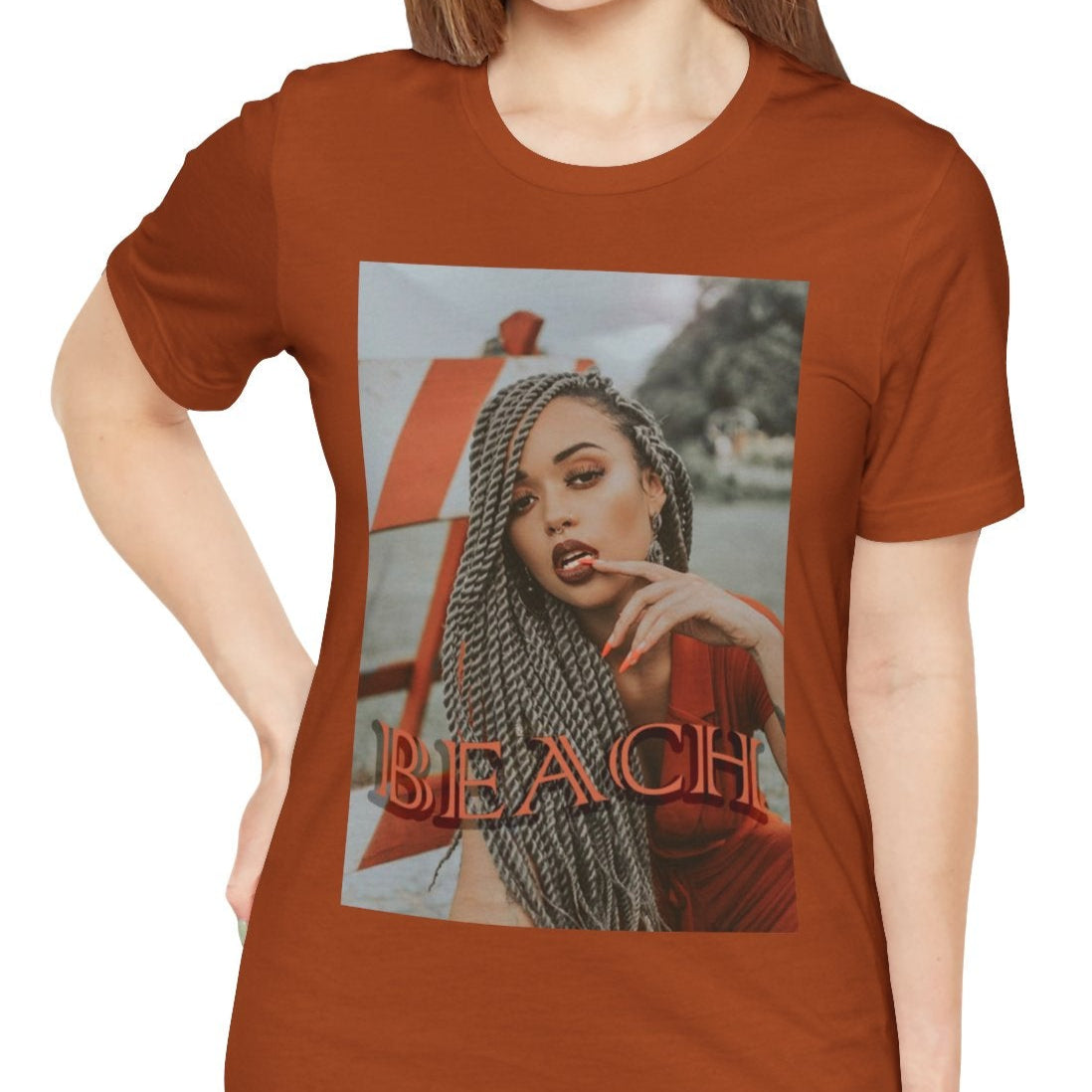 Portrait of a confident woman with long braids posing in front of an orange-and-white no-entry sign, printed on a unisex T-shirt in autumn, with the word “BEACH” in bold orange lettering. ModeK Vetements.
