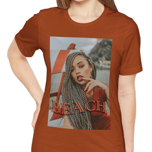 Portrait of a confident woman with long braids posing in front of an orange-and-white no-entry sign, printed on a unisex T-shirt in autumn, with the word “BEACH” in bold orange lettering. ModeK Vetements.