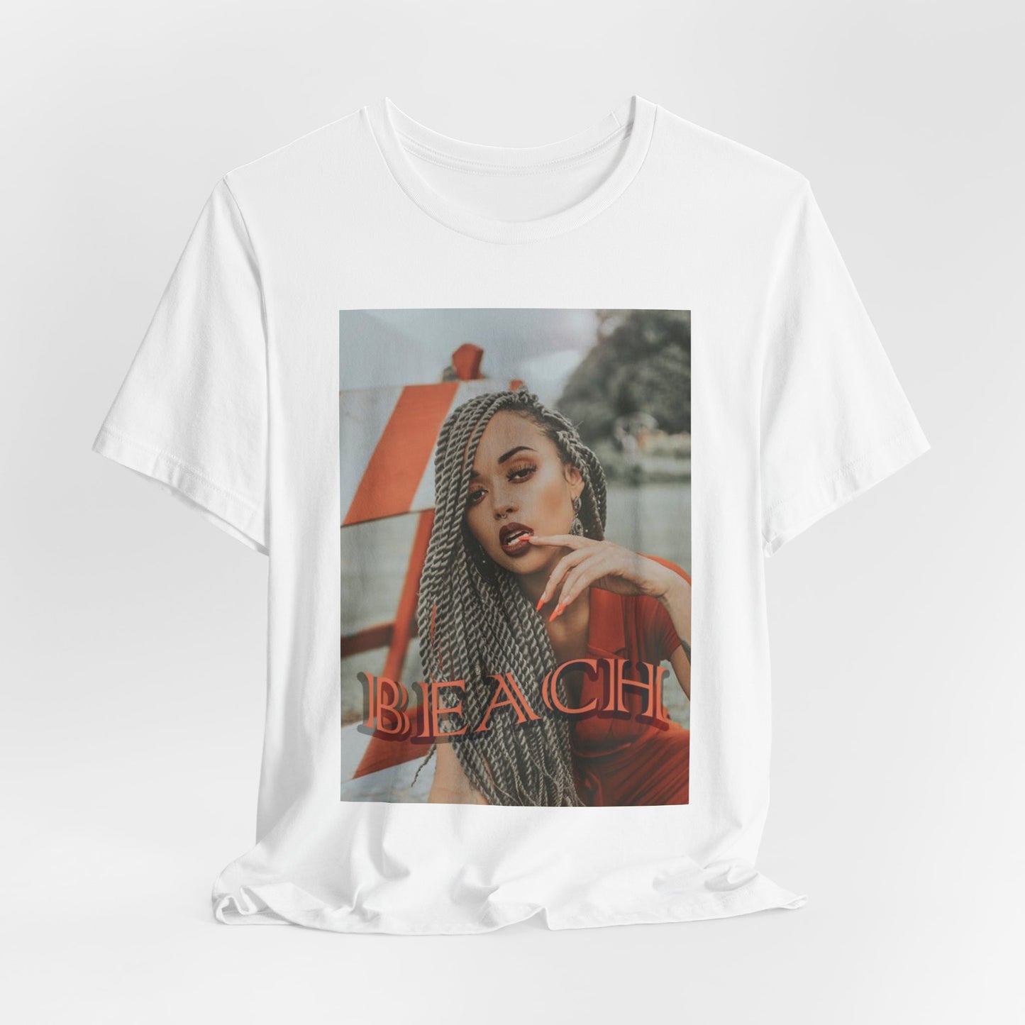 Portrait of a confident woman with long braids posing in front of an orange-and-white no-entry sign, printed on a unisex draped T-shirt in white, with the word “BEACH” in bold orange lettering. ModeK Vetements.