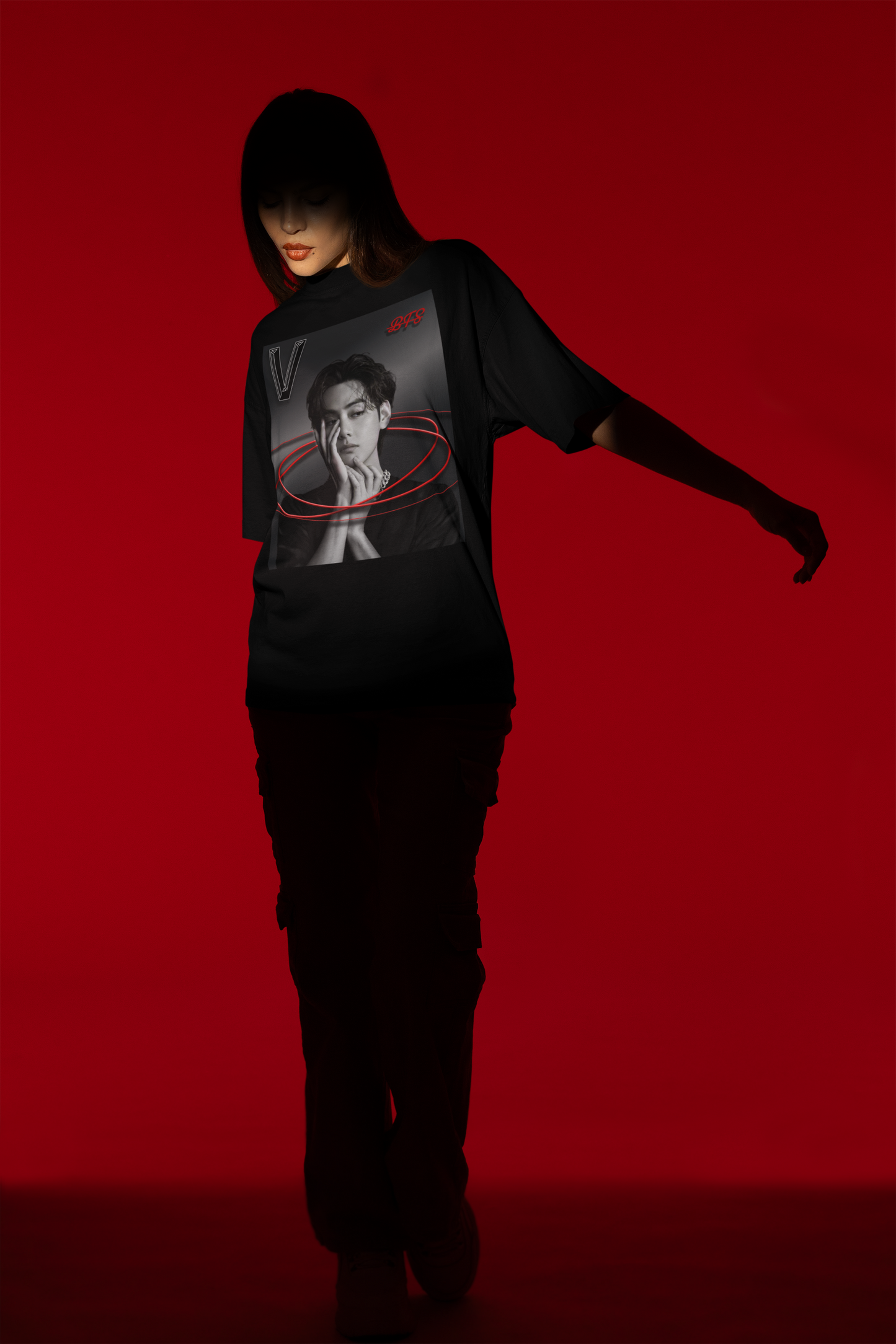 A full-size model stands in the spotlight over a red background, and showcases a unisex 100% cotton T-shirt in black. The front of the shirt displays a high-contrast black-and-white portrait of K-pop star V (Kim Taehyung) of BTS looking forward with a contemplative expression. Encircling him are vibrant red, glowing oval rings. A styled "V" appears in the top-left corner and a red "BTS" logo in the top-right corner. From ModeK Vetements.