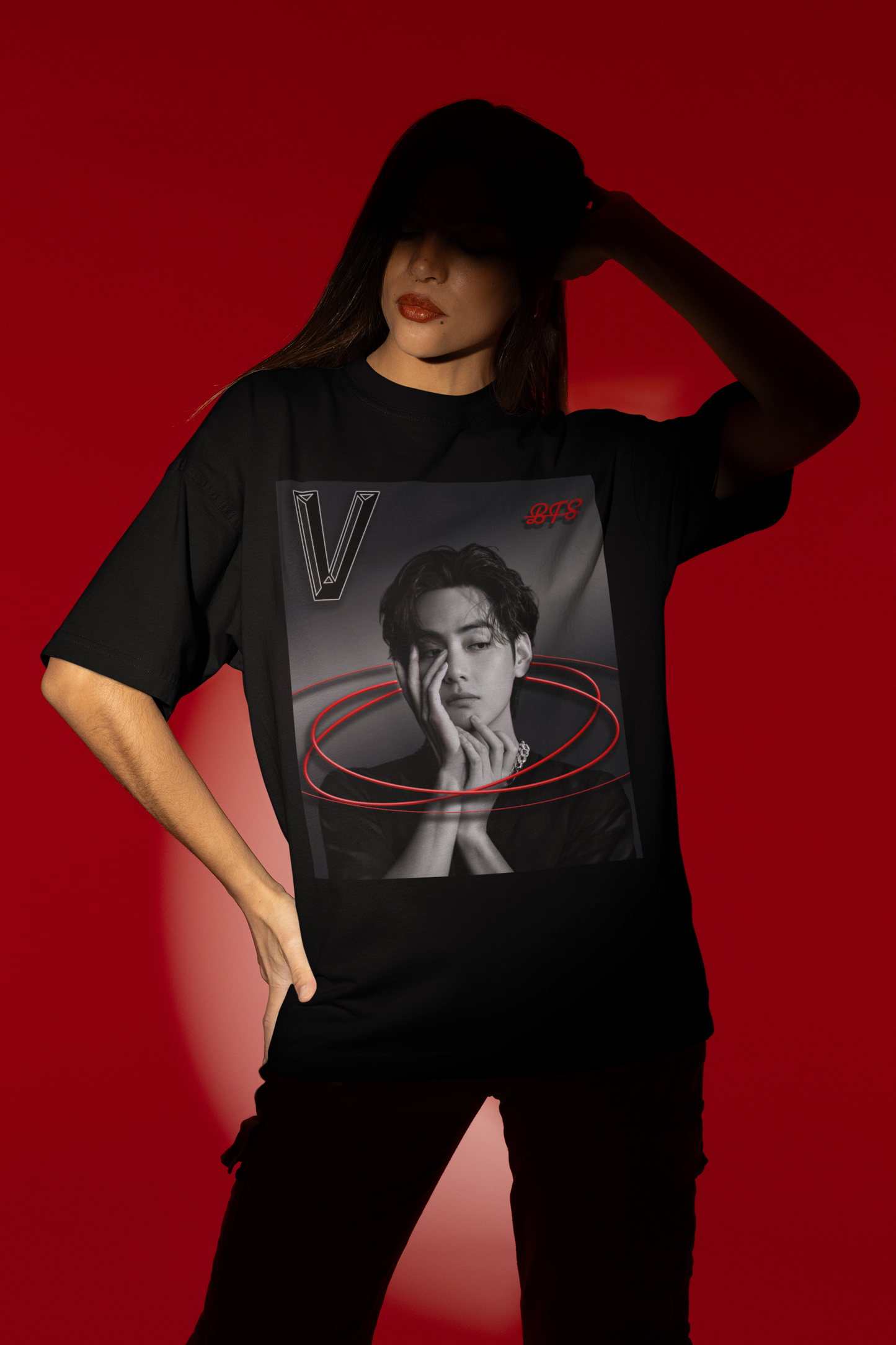 A model stands in the spotlight over a red background, with a hand in the hair, and showcases a unisex 100% cotton T-shirt in black. The front of the shirt displays a high-contrast black-and-white portrait of K-pop star V (Kim Taehyung) of BTS looking forward with a contemplative expression. Encircling him are vibrant red, glowing oval rings. A styled "V" appears in the top-left corner and a red "BTS" logo in the top-right corner. From ModeK Vetements.