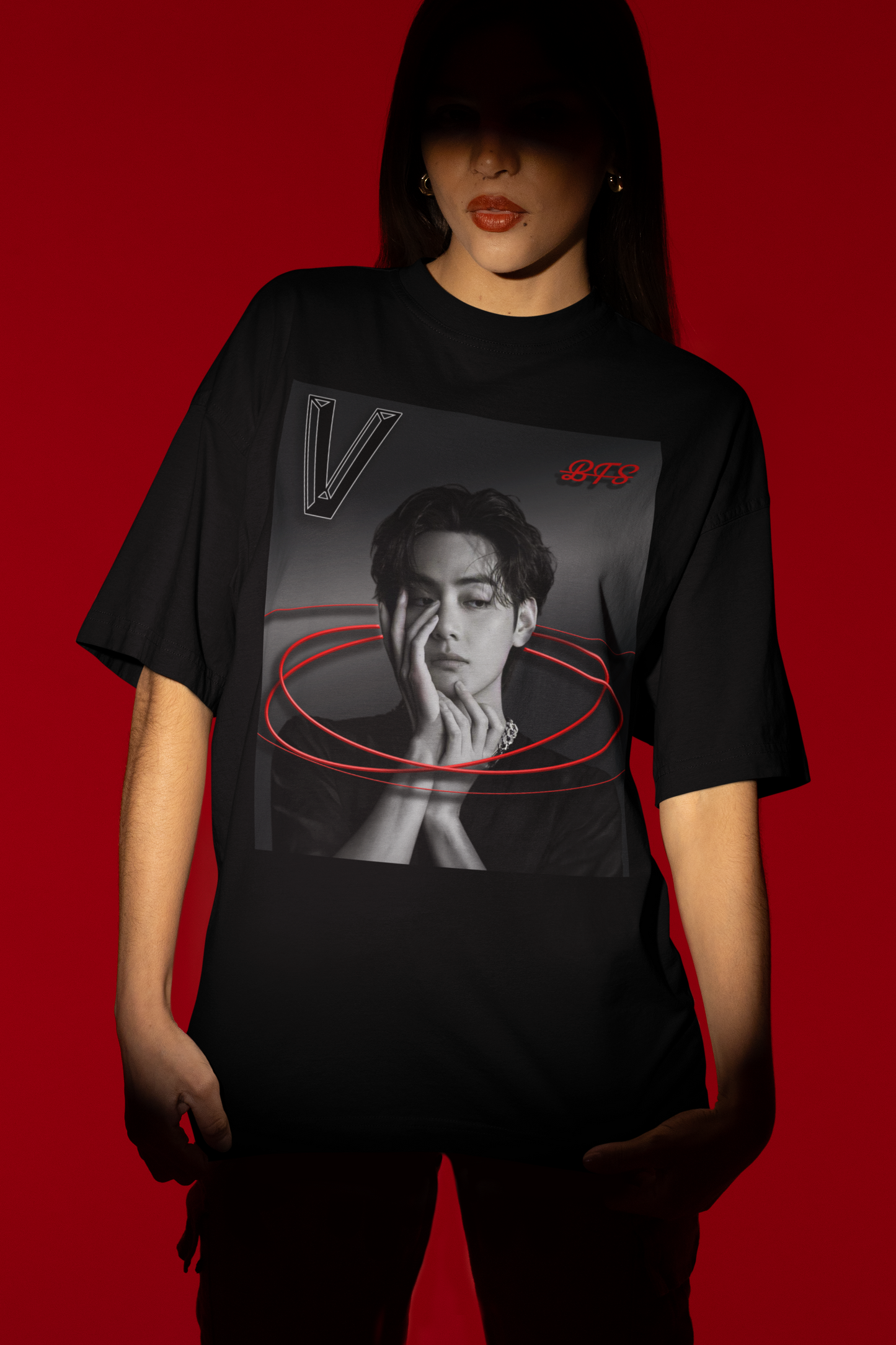 A model stands in the spotlight over a red background, and showcases a unisex 100% cotton T-shirt in black. The front of the shirt displays a high-contrast black-and-white portrait of K-pop star V (Kim Taehyung) of BTS looking forward with a contemplative expression. Encircling him are vibrant red, glowing oval rings. A styled "V" appears in the top-left corner and a red "BTS" logo in the top-right corner. From ModeK Vetements.