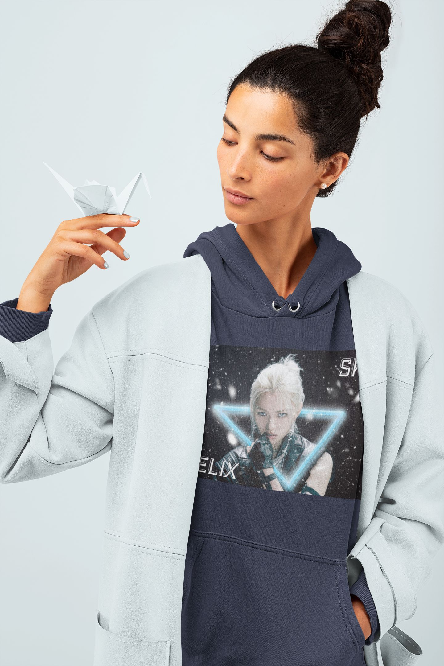 Model holding an origami and showcasing the Unisex cotton-poly blend hoodie in navy, featuring a visual print of Felix from Stray Kids, framed by a glowing blue neon triangle amid falling snow on a dark background. From ModeK Vetements.