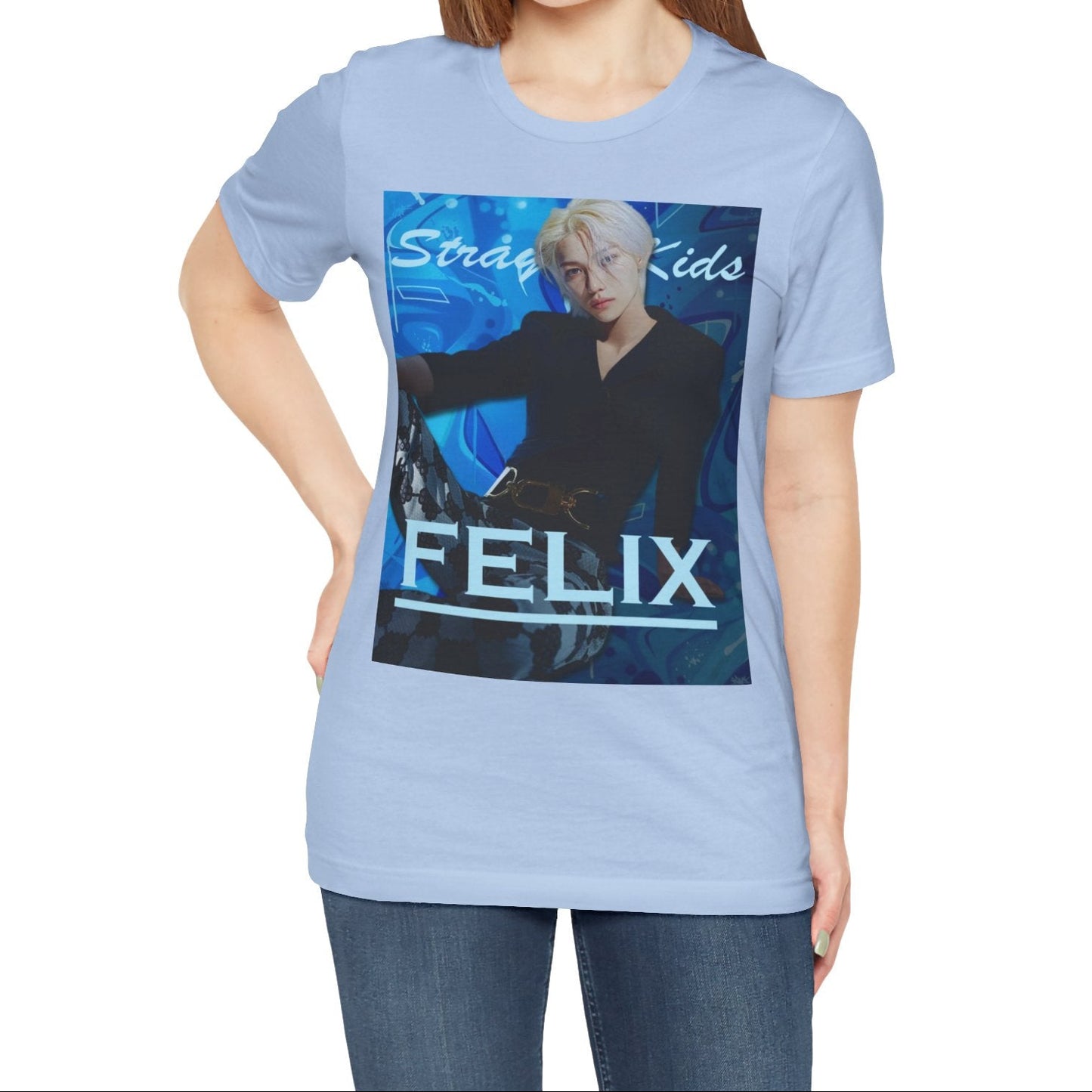 Female model showcasing the unisex, 100% airlume combed & ring-spun cotton t-shirt in baby blue, featuring a graphic design. It shows Felix from Stray Kids in a provocative pose, wearing a structured black suit jacket and oriental-style patterned pants. The background is a multi-toned blue graffiti. From ModeK Vetements.