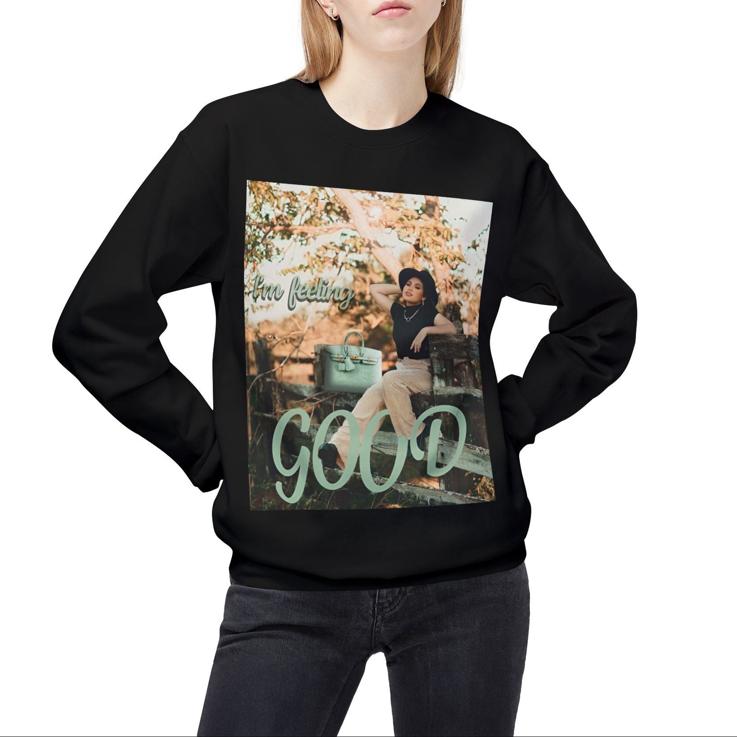 Model wearing a unisex sweatshirt in black featuring a photo of a woman in beige trousers and black top on a rustic fence. The gum leaf green “I’m feeling GOOD” text echoes the handbag and wraps visually around her legs. — From ModeK Vetements