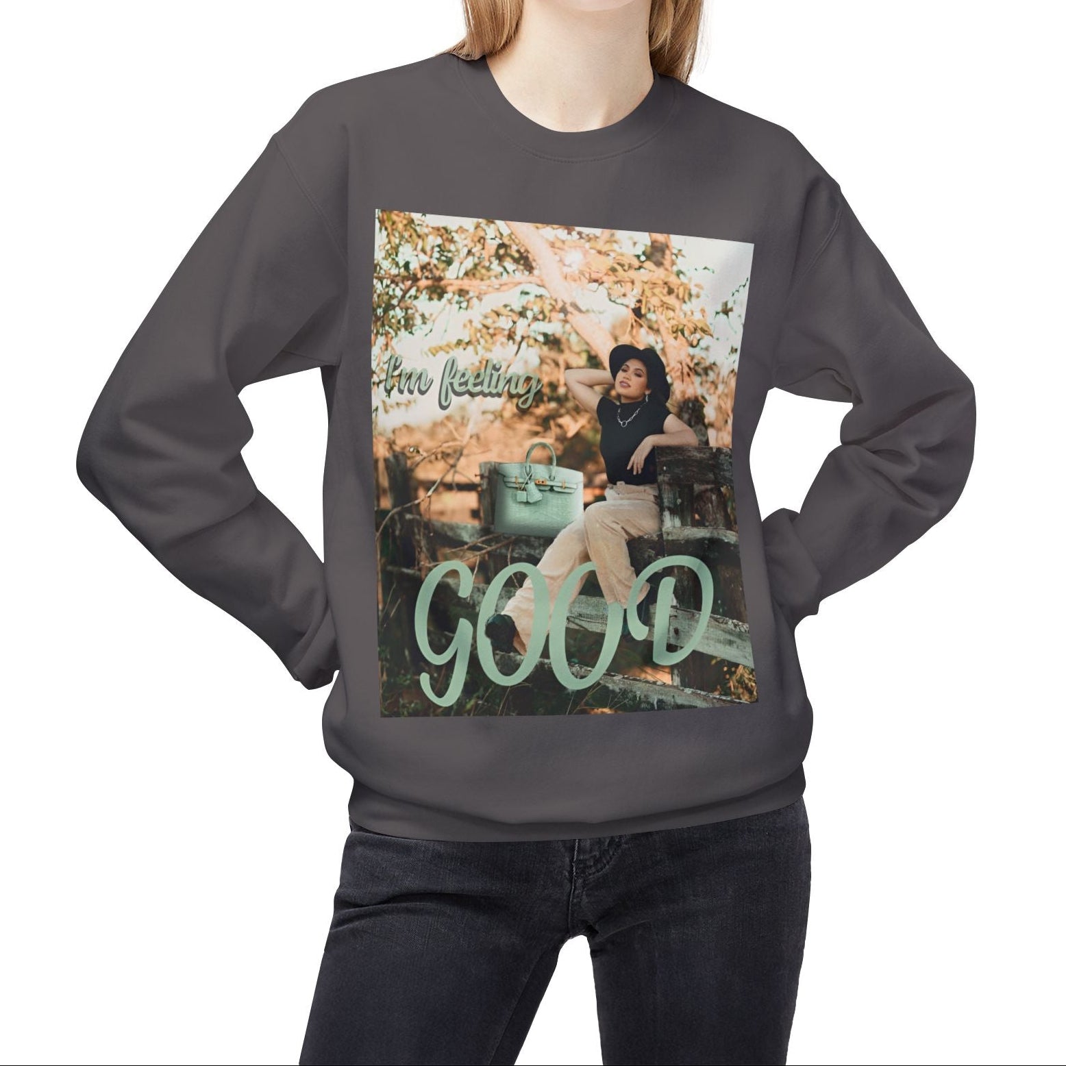 Model wearing a unisex sweatshirt in charcoal featuring a photo of a woman in beige trousers and black top on a rustic fence. The gum leaf green “I’m feeling GOOD” text echoes the handbag and wraps visually around her legs. — From ModeK Vetements