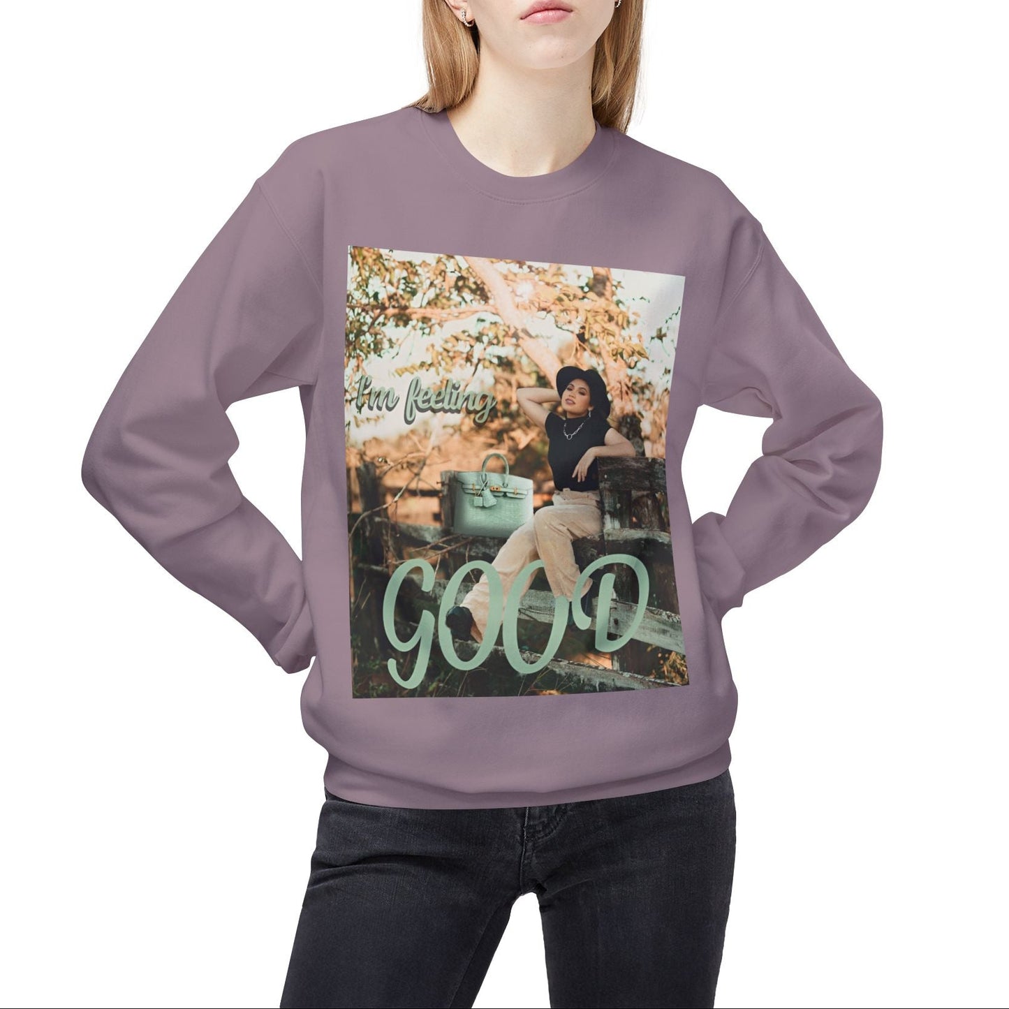 Model wearing a unisex sweatshirt in paragon featuring a photo of a woman in beige trousers and black top on a rustic fence. The gum leaf green “I’m feeling GOOD” text echoes the handbag and wraps visually around her legs. — From ModeK Vetements