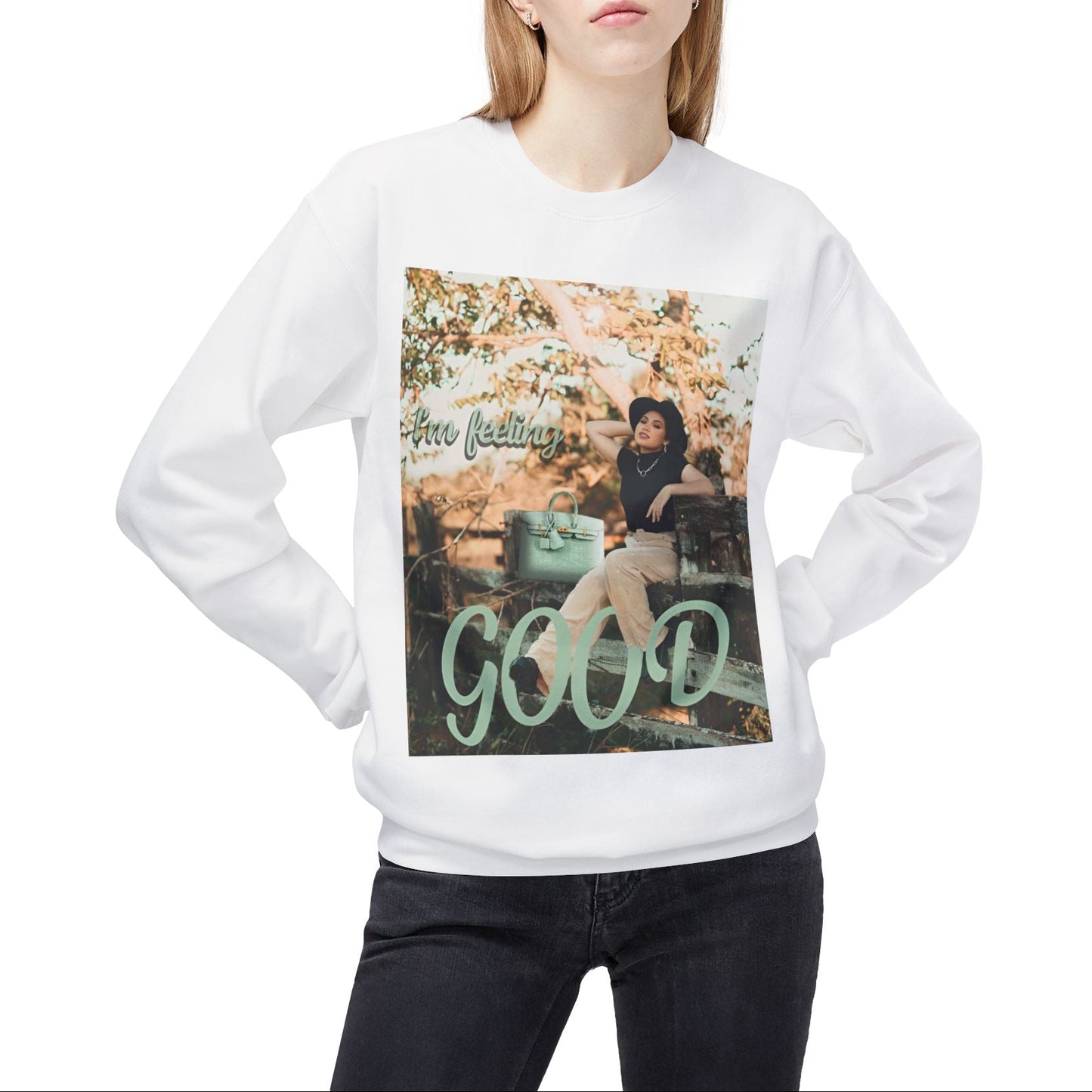 Model wearing a unisex sweatshirt in white featuring a photo of a woman in beige trousers and black top on a rustic fence. The gum leaf green “I’m feeling GOOD” text echoes the handbag and wraps visually around her legs. — From ModeK Vetements