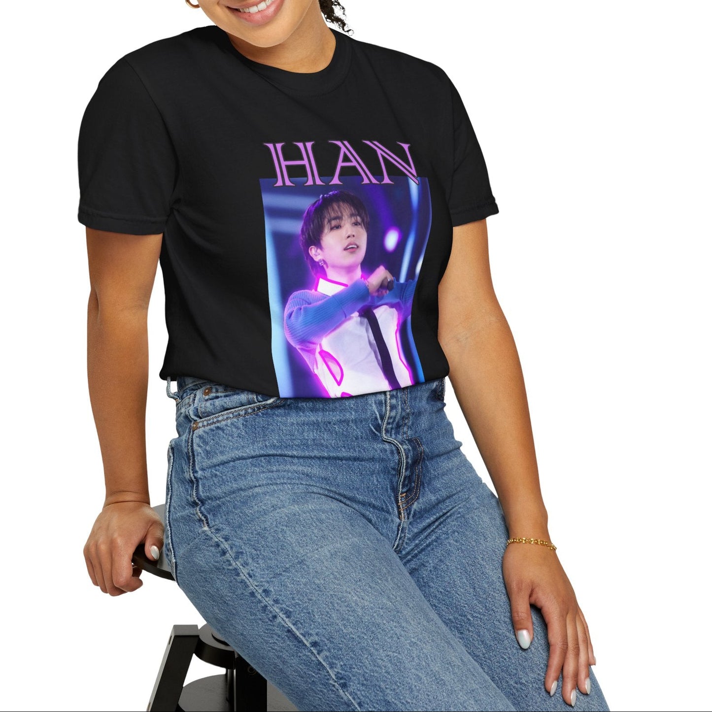 Model sitting on a stool and showcasing the Han Stray Kids Unisex T-Shirt in black – Neon Energy. A striking design featuring Han performing on stage, outlined in bold pink neon lights. The glowing effect adds a futuristic, high-energy feel, capturing his electrifying stage presence. 100% ring-spun cotton from ModeK Vetements.