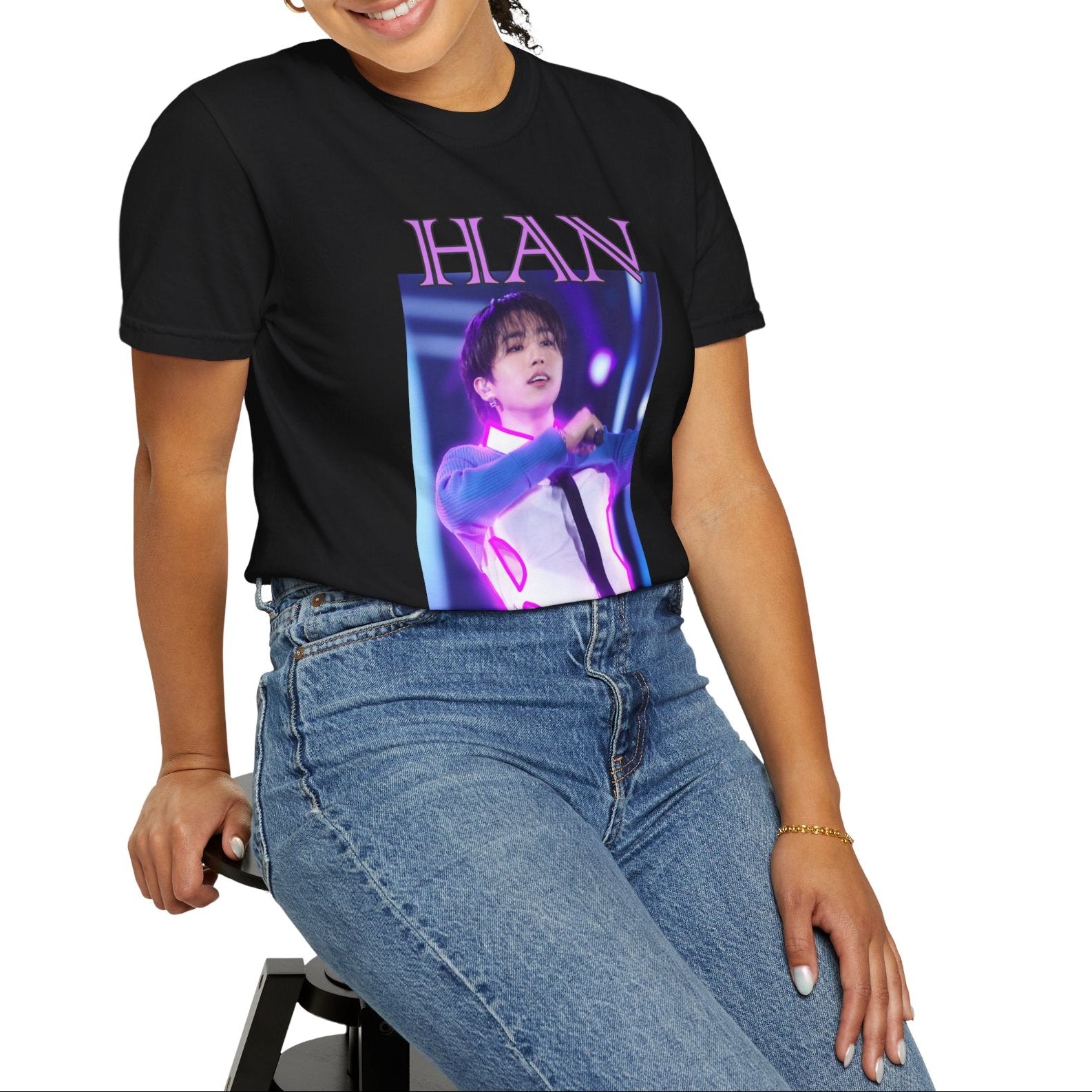 Model sitting on a stool and showcasing the Han Stray Kids Unisex T-Shirt in black – Neon Energy. A striking design featuring Han performing on stage, outlined in bold pink neon lights. The glowing effect adds a futuristic, high-energy feel, capturing his electrifying stage presence. 100% ring-spun cotton from ModeK Vetements.
