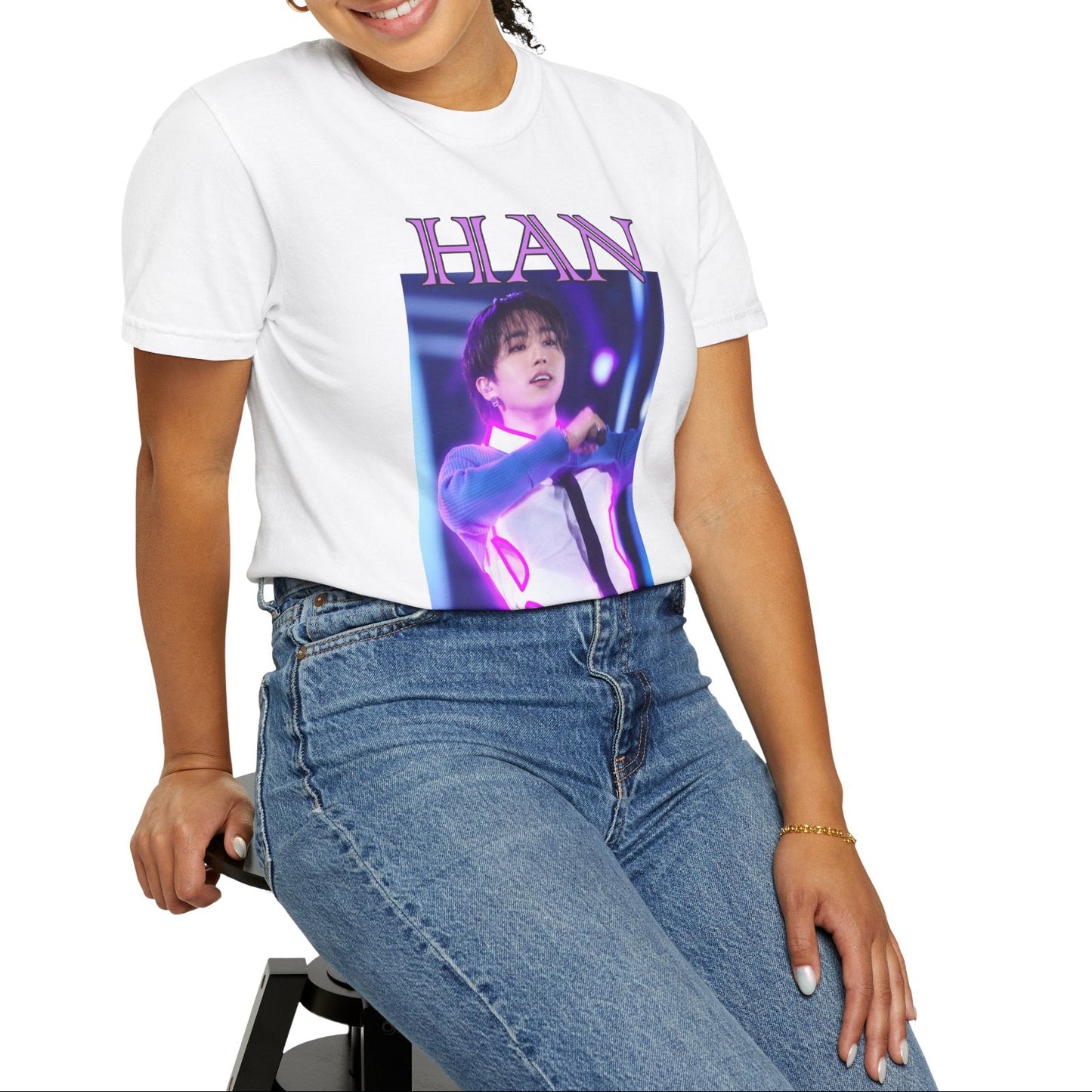 Model sitting on a stool and showcasing the Han Stray Kids Unisex T-Shirt in white – Neon Energy. A striking design featuring Han performing on stage, outlined in bold pink neon lights. The glowing effect adds a futuristic, high-energy feel, capturing his electrifying stage presence. 100% ring-spun cotton from ModeK Vetements.