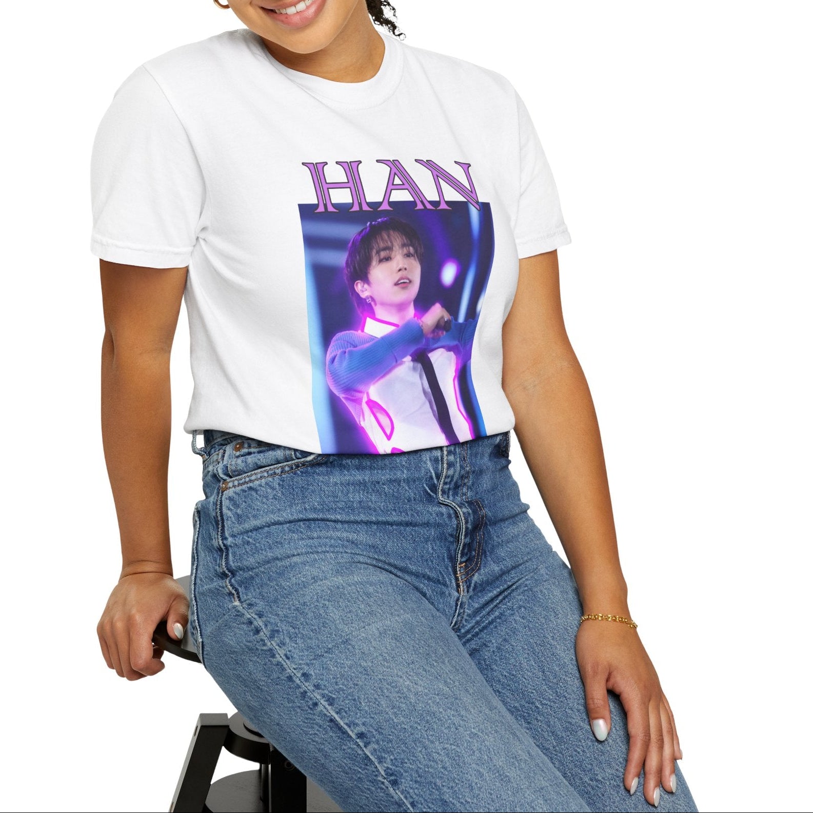 Model sitting on a stool and showcasing the Han Stray Kids Unisex T-Shirt in white – Neon Energy. A striking design featuring Han performing on stage, outlined in bold pink neon lights. The glowing effect adds a futuristic, high-energy feel, capturing his electrifying stage presence. 100% ring-spun cotton from ModeK Vetements.