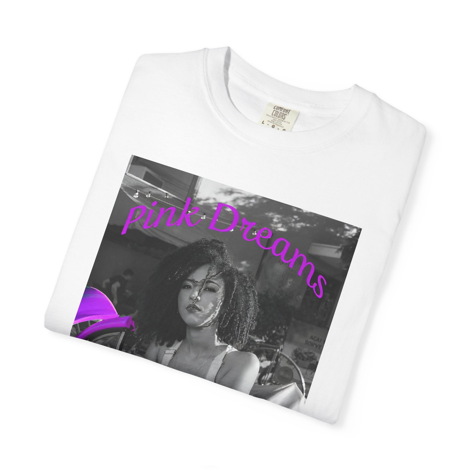 Folded Unisex T-shirt in white, made from 100% ring-spun cotton featuring a printed design where a pinkish purple race car dominates a monochrome scene, with a girl leaning calmly against it. The words “Pink Dreams” curve above, evoking attitude, stillness, and the feeling of living on her own terms. From ModeK Vetements.