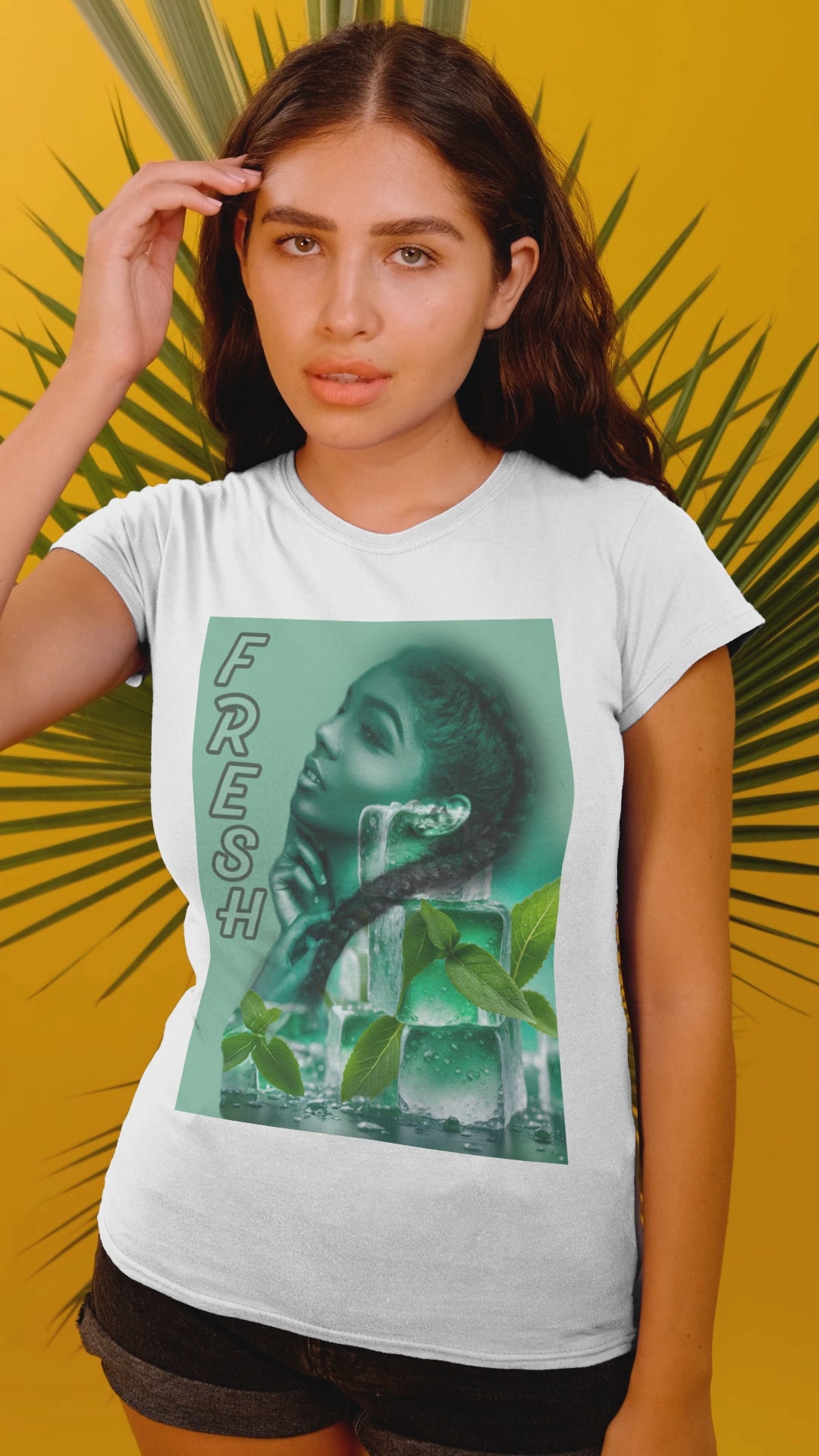 Video showcasing the Unisex cotton t-shirt in white, featuring a layered image of a woman behind translucent ice cubes with mint leaves. Her braid crosses in front of the cubes. Vertical “FRESH” lettering is outlined but empty, adding a floating, airy effect. Turquoise background. Printed on 100% US cotton, ethically made and Oeko-Tex certified. ModeK Vetements.