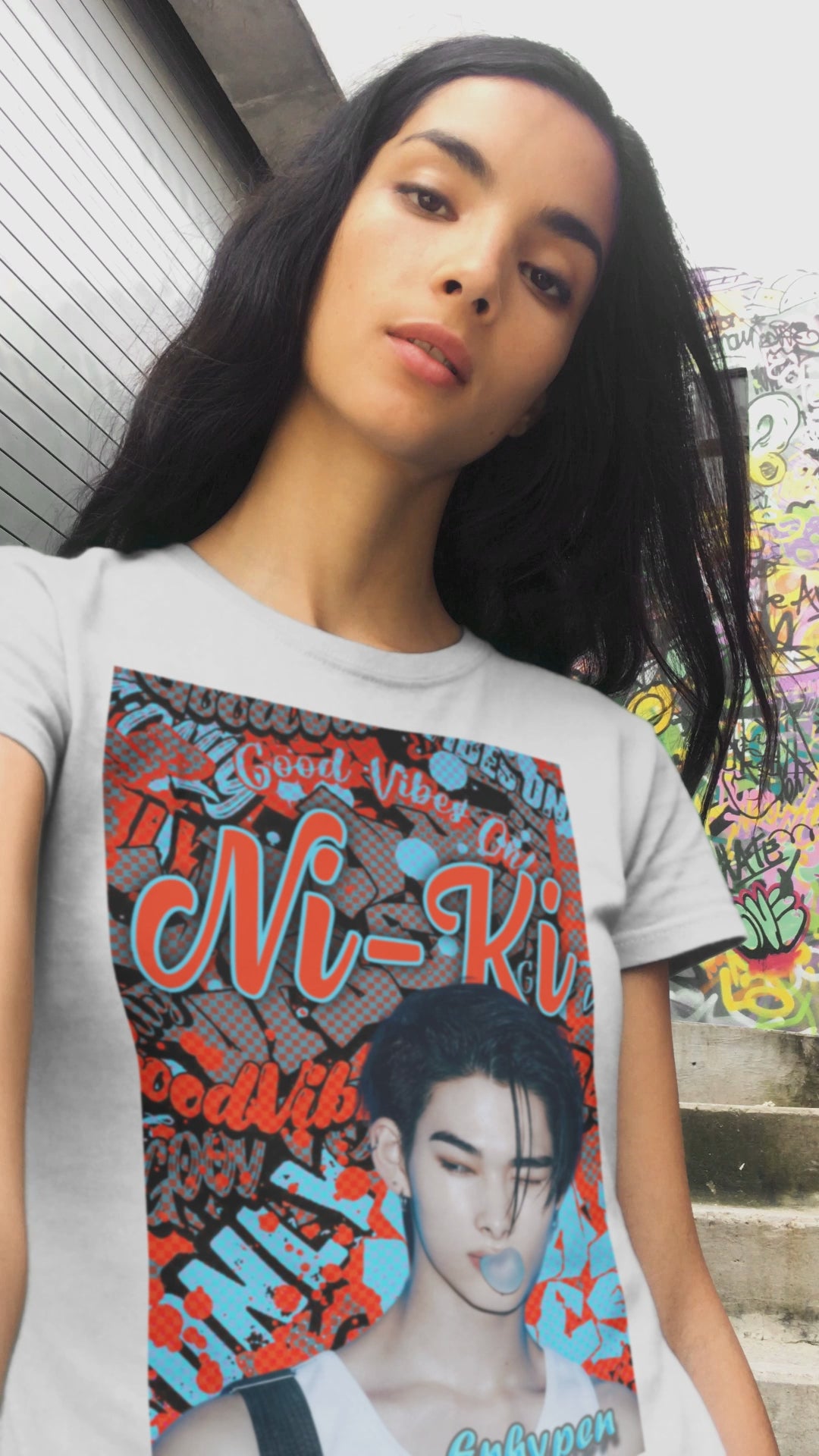 Video showcasing the Unisex airlume cotton T-shirt in white, featuring Ni-Ki of ENHYPEN layered into a bold graffiti-style design with Rosso Corsa red and Malibu Blue. The layout includes digital splashes, checkerboard effects, and a bubble gum moment frozen mid-pop. From ModeK Vetements.