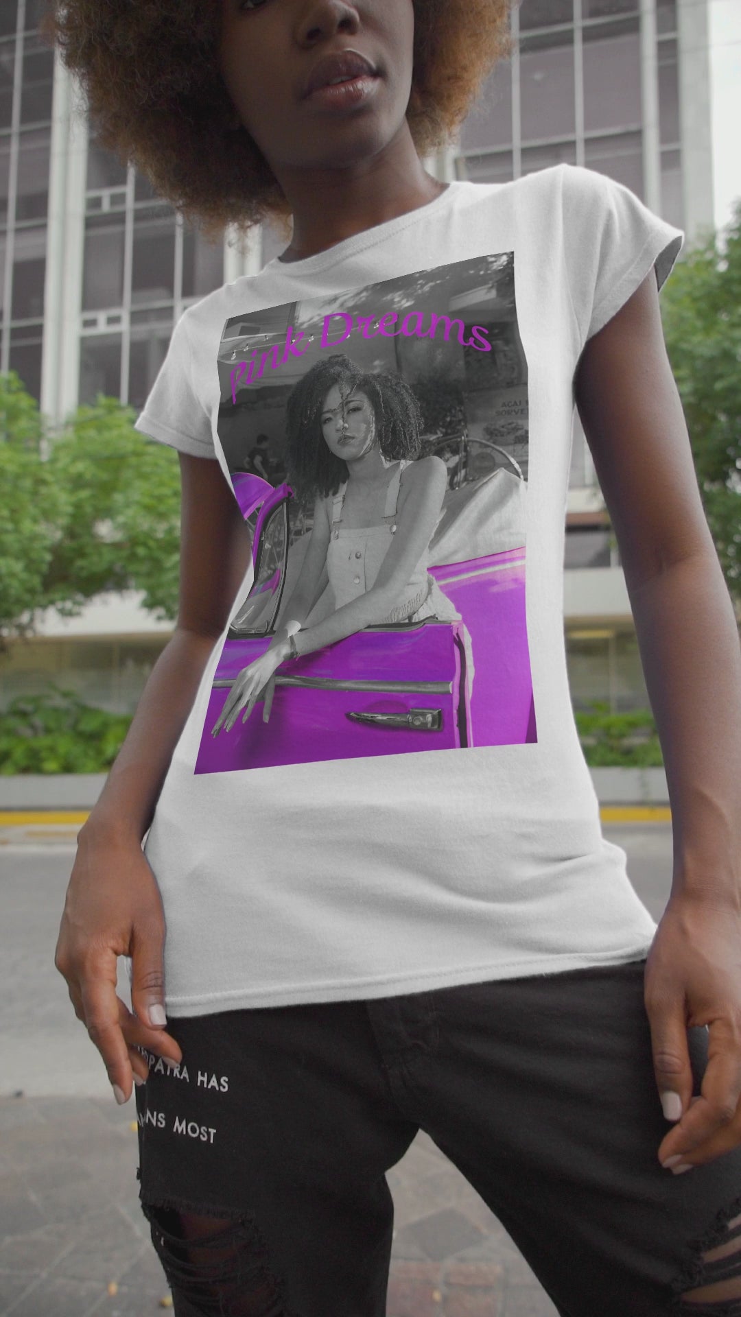 Video showcasing the Unisex T-shirt in white, made from 100% ring-spun cotton featuring a printed design where a pinkish purple race car dominates a monochrome scene, with a girl leaning calmly against it. The words “Pink Dreams” curve above, evoking attitude, stillness, and the feeling of living on her own terms. From ModeK Vetements.