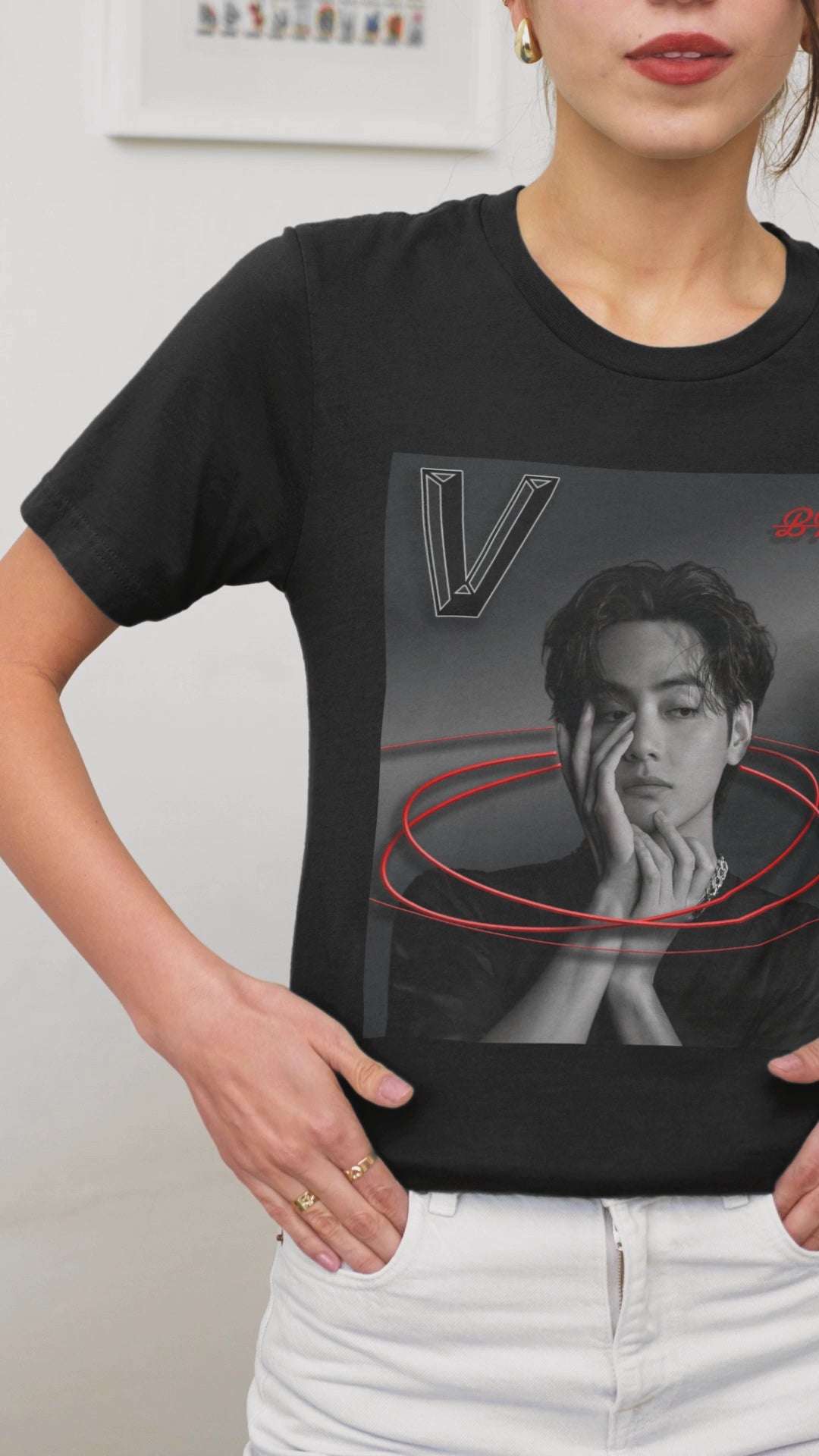 Video showcasing the unisex 100% cotton T-shirt in black. The front of the shirt displays a high-contrast black-and-white portrait of K-pop star V (Kim Taehyung) of BTS looking forward with a contemplative expression. Encircling him are vibrant red, glowing oval rings. A styled "V" appears in the top-left corner and a red "BTS" logo in the top-right corner. From ModeK Vetements.
