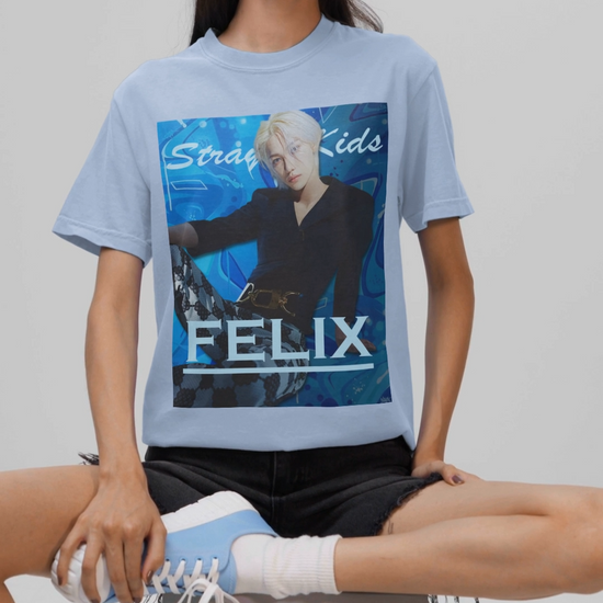 Video showcasing the unisex, 100% airlume combed & ring-spun cotton t-shirt in baby blue, featuring a graphic design. It shows Felix from Stray Kids in a provocative pose, wearing a structured black suit jacket and oriental-style patterned pants. The background is a multi-toned blue graffiti. From ModeK Vetements.