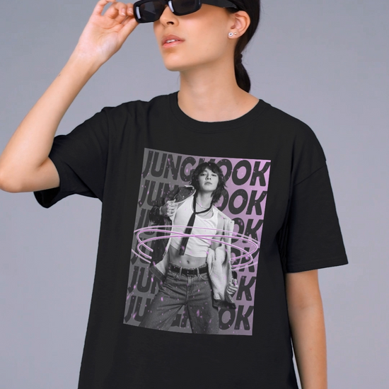 Video showcasing the unisex 100% ring-spun cotton T-Shirt in black, featuring BTS's Jungkook. He is posing against a dark background with "JUNGKOOK" repeated in large black typography and a charcoal-lavender gradient with pink paint splatters. Three glowing, thin magenta hoops encircle his waist, creating a sense of motion, making him even more provocative. From ModeK Vetements.