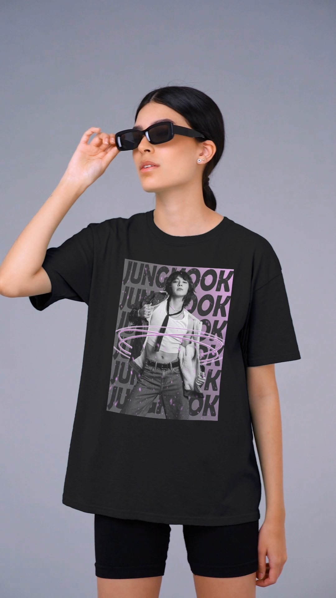 Video showcasing the unisex 100% ring-spun cotton T-Shirt in black, featuring BTS's Jungkook. He is posing against a dark background with "JUNGKOOK" repeated in large black typography and a charcoal-lavender gradient with pink paint splatters. Three glowing, thin magenta hoops encircle his waist, creating a sense of motion, making him even more provocative. From ModeK Vetements.