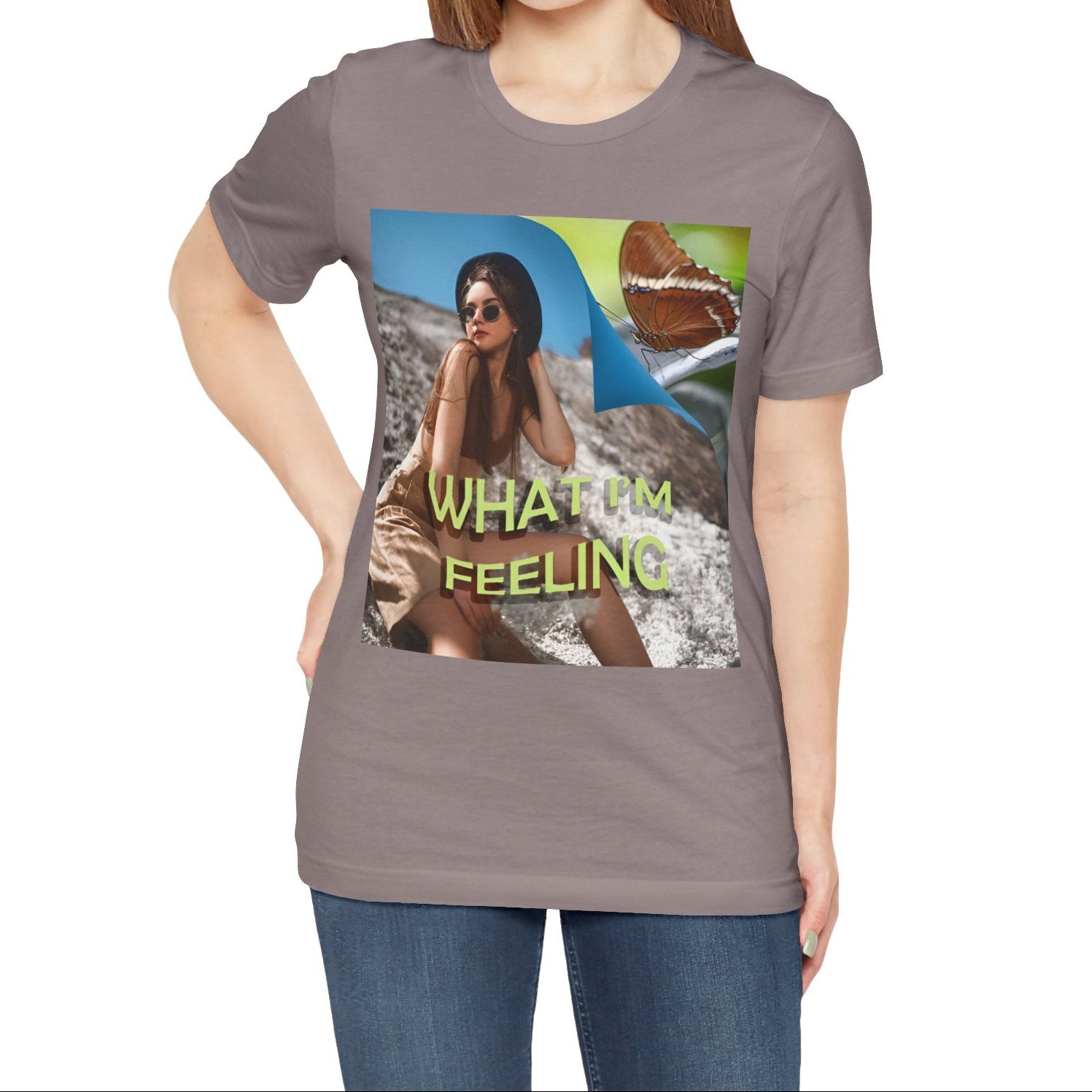 Unisex jersey T-Shirt in pebble brown featuring a woman sitting on a sunlit rocky beach with mint text “WHAT I’M FEELING.” A folded corner reveals a rusty-tipped page butterfly. — From ModeK Vetements.
