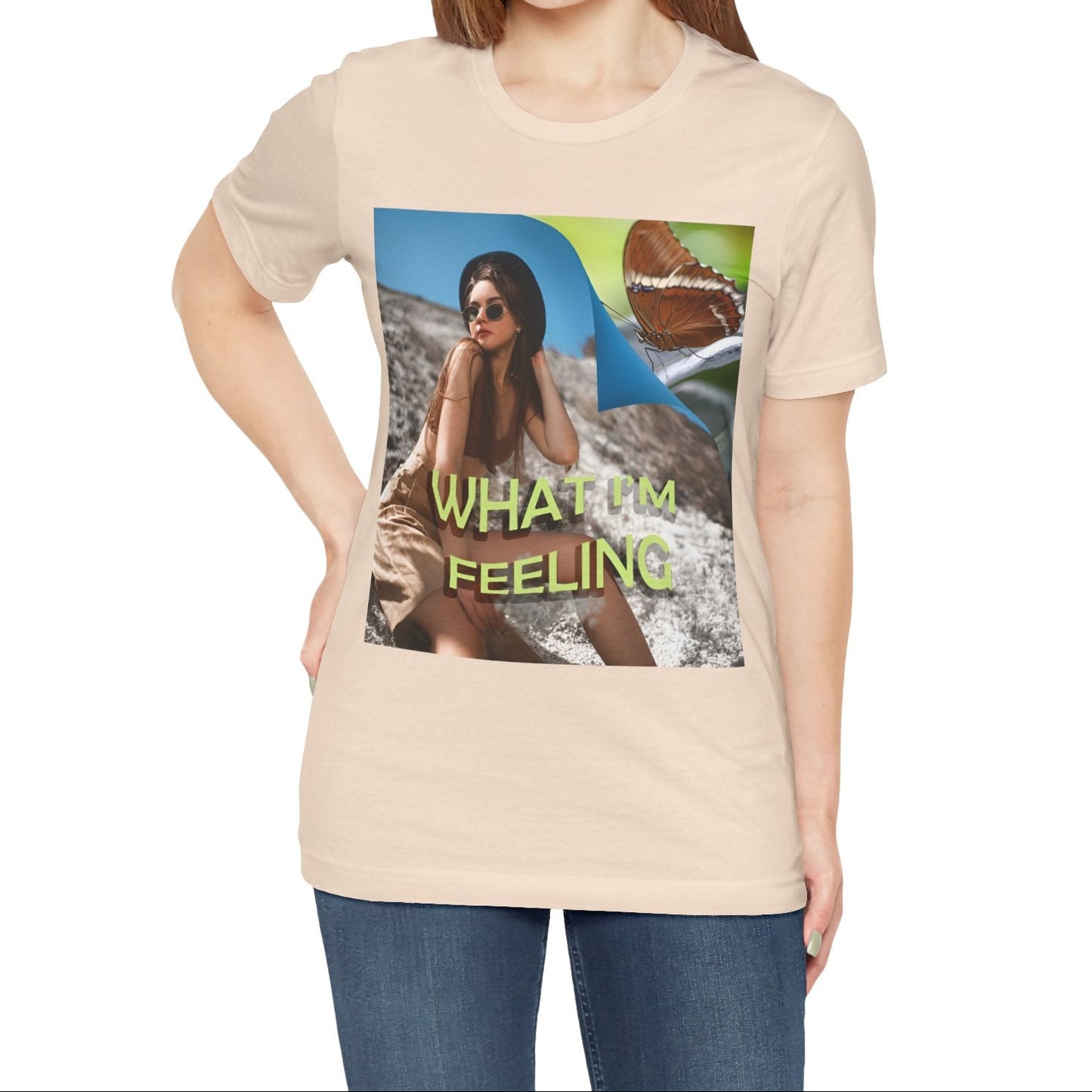 Unisex jersey T-Shirt in soft cream featuring a woman sitting on a sunlit rocky beach with mint text “WHAT I’M FEELING.” A folded corner reveals a rusty-tipped page butterfly. — From ModeK Vetements.