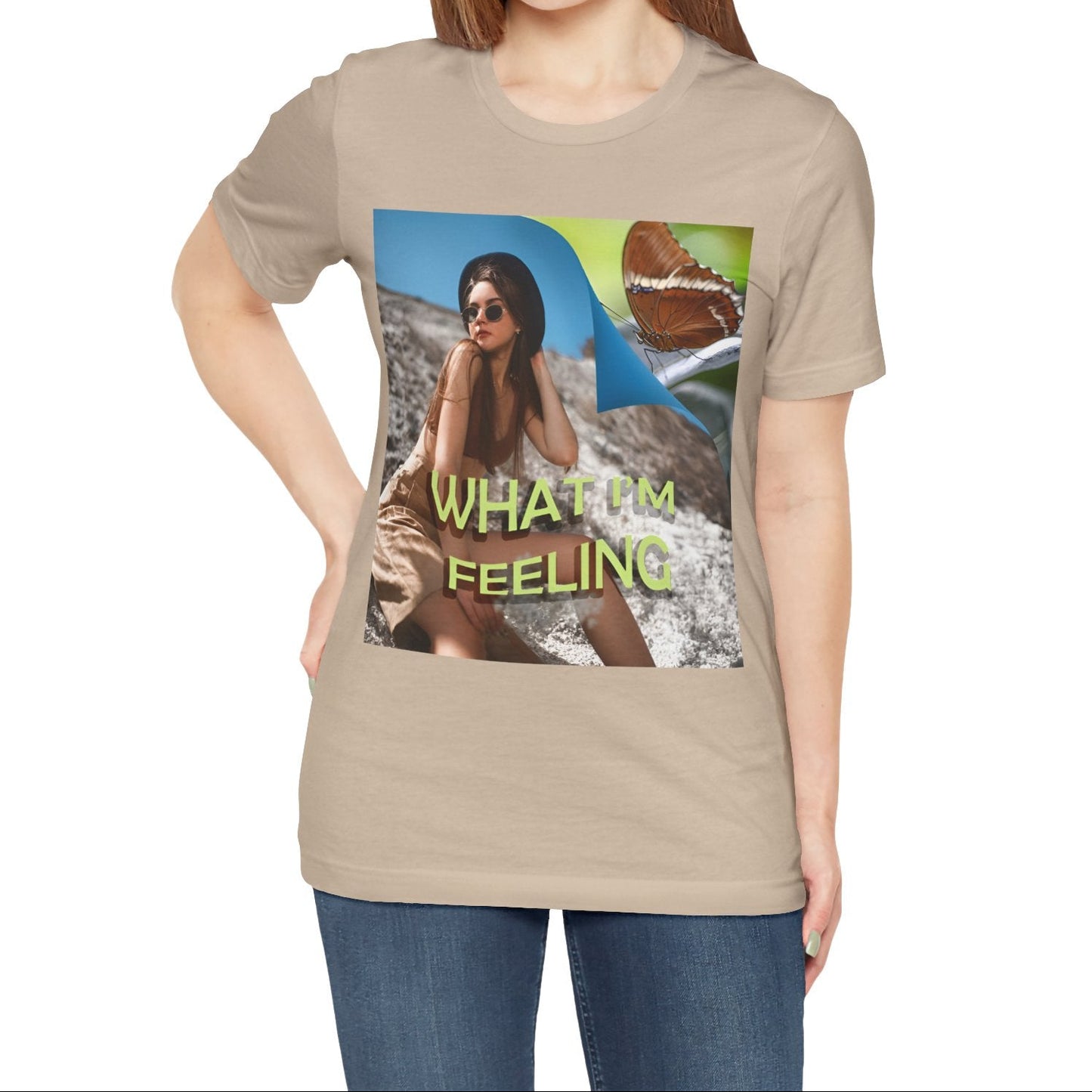 Unisex jersey T-Shirt in tan featuring a woman sitting on a sunlit rocky beach with mint text “WHAT I’M FEELING.” A folded corner reveals a rusty-tipped page butterfly. — From ModeK Vetements.
