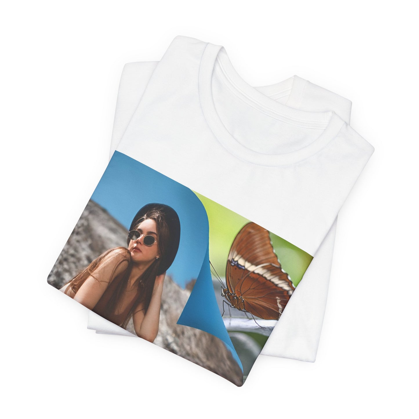 Folded unisex jersey T-Shirt in white featuring a woman sitting on a sunlit rocky beach with mint text “WHAT I’M FEELING.” A folded corner reveals a rusty-tipped page butterfly. — From ModeK Vetements.