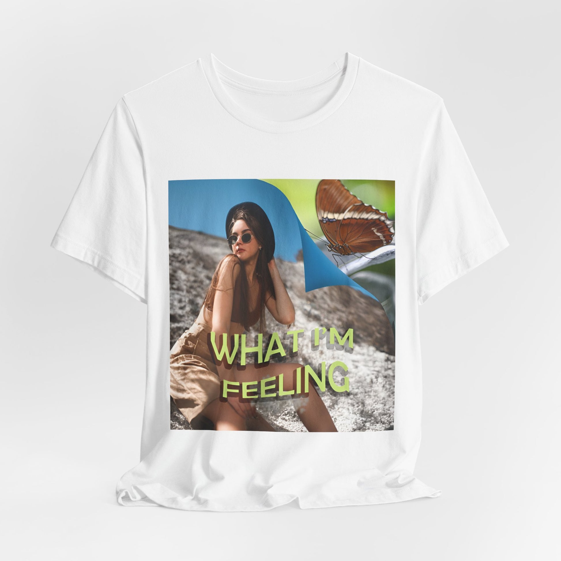 Hanging unisex jersey T-Shirt in white featuring a woman sitting on a sunlit rocky beach with mint text “WHAT I’M FEELING.” A folded corner reveals a rusty-tipped page butterfly. — From ModeK Vetements.