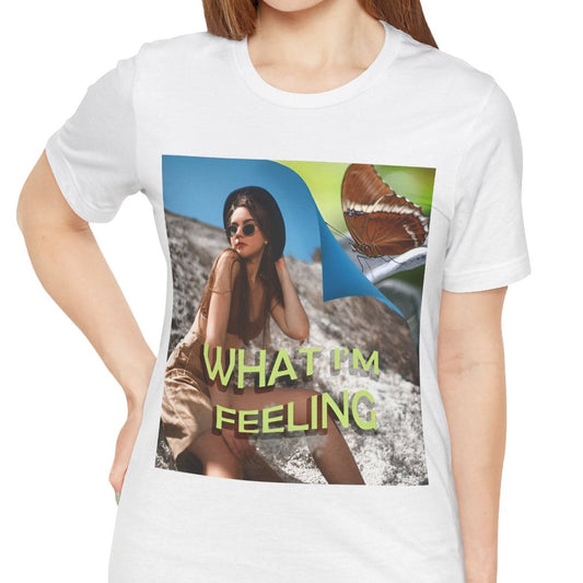 Unisex jersey T-Shirt in white featuring a woman sitting on a sunlit rocky beach with mint text “WHAT I’M FEELING.” A folded corner reveals a rusty-tipped page butterfly. — From ModeK Vetements.