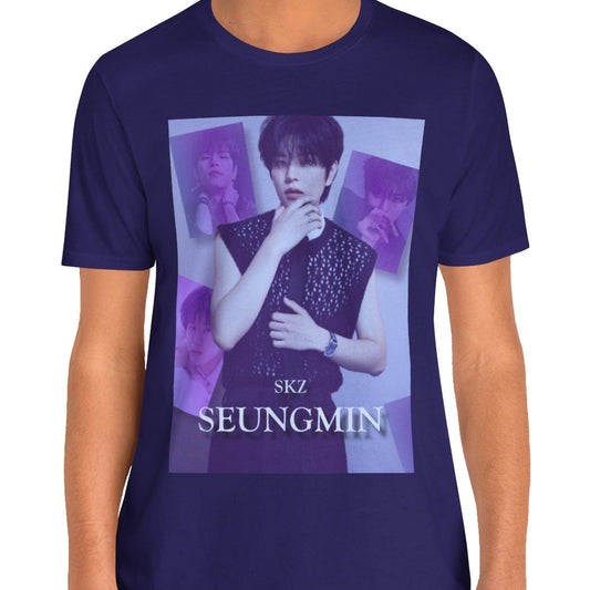 Male model showcasing the unisex, jersey, airlume combed and ringspun cotton T-Shirt in team navy, with a graphic design. It features Stray Kids' Seungmin in a sleeveless crochet top and blueberry tie. He poses thoughtfully against a periwinkle backdrop with a mist-covered collage of lavender, magazine-style photos. At the bottom, "SKZ SEUNGMIN" is written in pale lily-blue with a soft drop shadow. From ModeK Vetments.