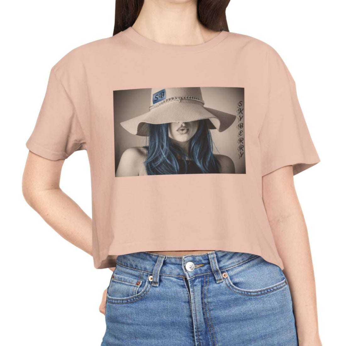 Design printed on a women’s crop tee in milk berry, made from 100% combed cotton. It features a stylized woman with soft blue hair and a wide felt hat, her gaze hidden. Above her, the word “Skyberry” appears in black lettering. The composition evokes quiet mystery and the feeling of a summer mirage. From Modek Vetements.