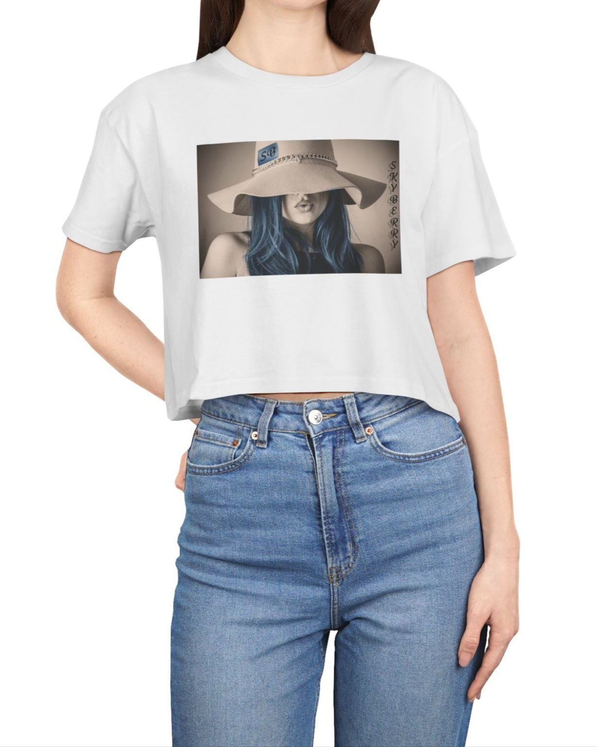 Design printed on a women’s crop tee in white, made from 100% combed cotton. It features a stylized woman with soft blue hair and a wide felt hat, her gaze hidden. Above her, the word “Skyberry” appears in black lettering. The composition evokes quiet mystery and the feeling of a summer mirage. From Modek Vetements.