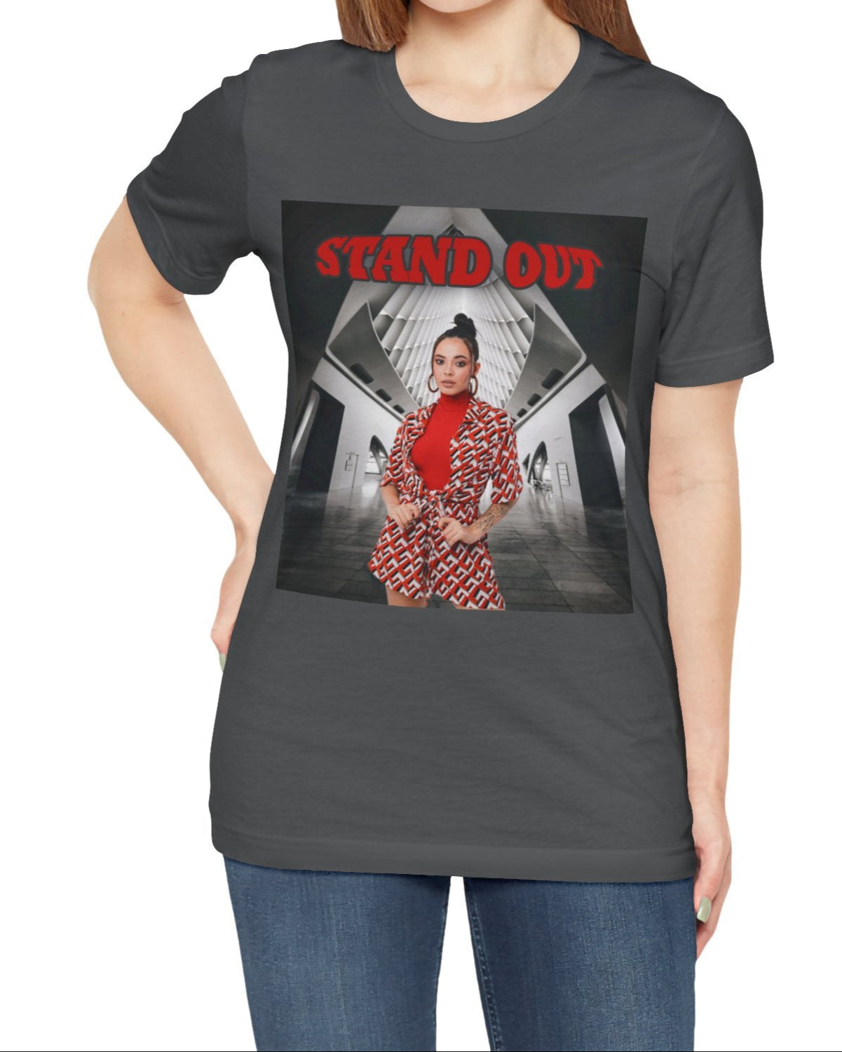 Unisex airlume cotton T-Shirt in asphalt, featuring a Woman in bold red geometric outfit standing confidently in a futuristic black-and-white architectural space. The "STAND OUT" in striking retro-style block font above her is a visual statement of individuality. From ModeK Vetements.