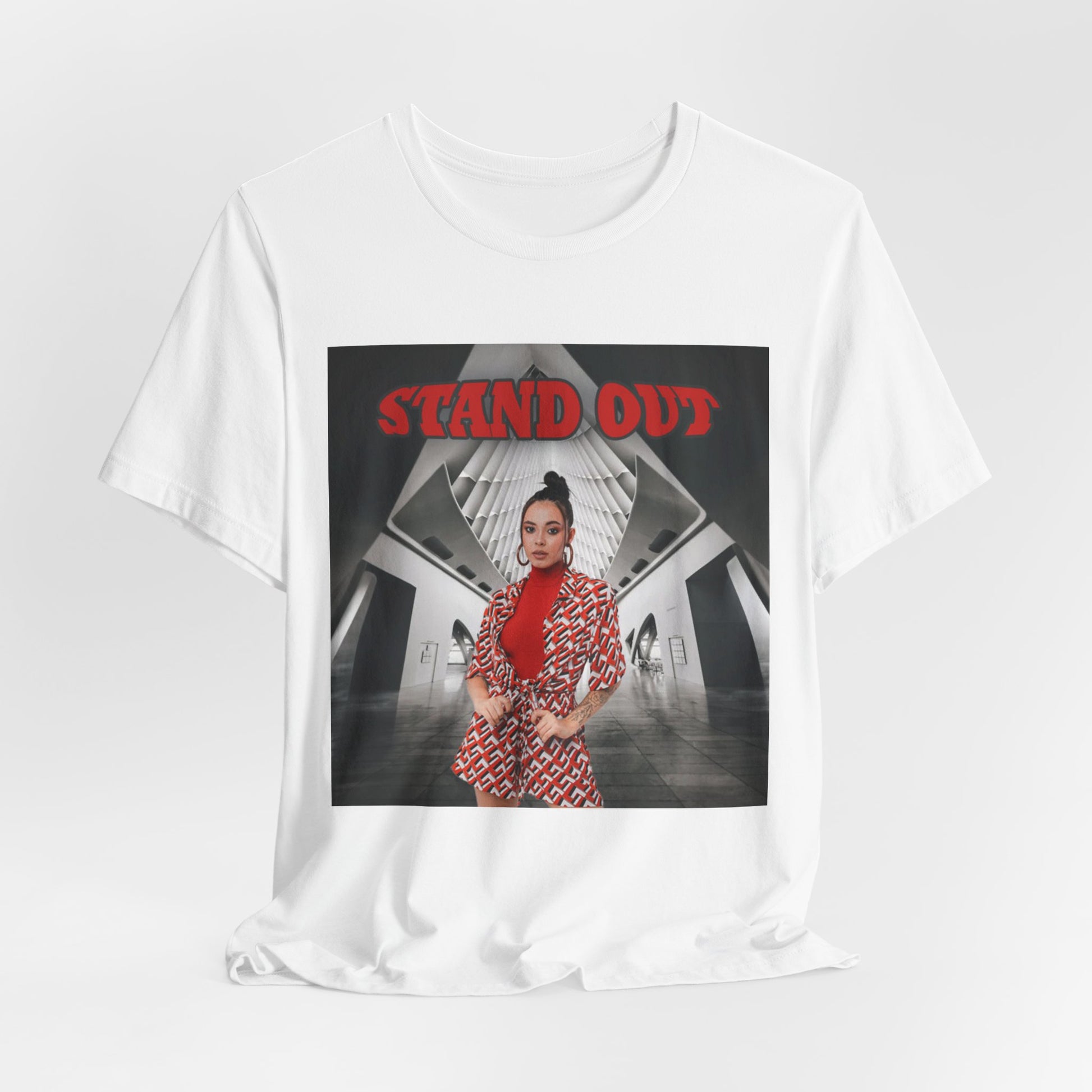 Draped Unisex cotton T-Shirt in white, featuring a Woman in bold red geometric outfit standing confidently in a futuristic black-and-white architectural space. The "STAND OUT" in striking retro-style block font above her is a visual statement of individuality. From ModeK Vetements.