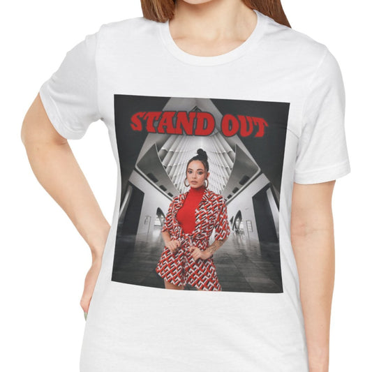 Unisex airlume cotton T-Shirt in white, featuring a Woman in bold red geometric outfit standing confidently in a futuristic black-and-white architectural space. The "STAND OUT" in striking retro-style block font above her is a visual statement of individuality. From ModeK Vetements.