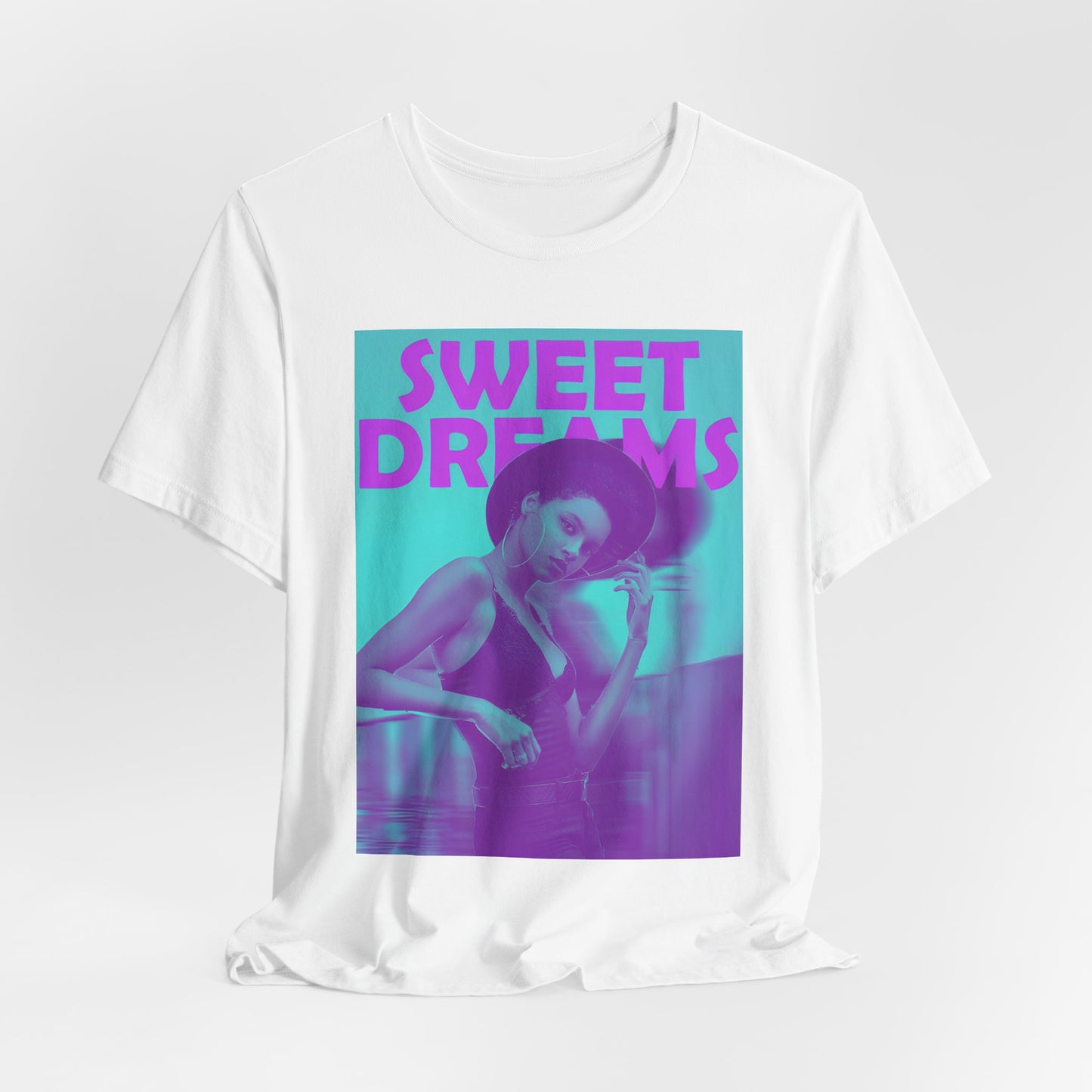 Draped unisex cotton t-shirt in white, featuring a woman in a wide-brimmed hat appearing clearly over a blurred motion version of herself, as if caught between dream and sleep. The lilac-pink words “SWEET DREAMS” float above on a medium turquoise background, evoking the gentle melancholy of May evenings. From ModeK Vetements.