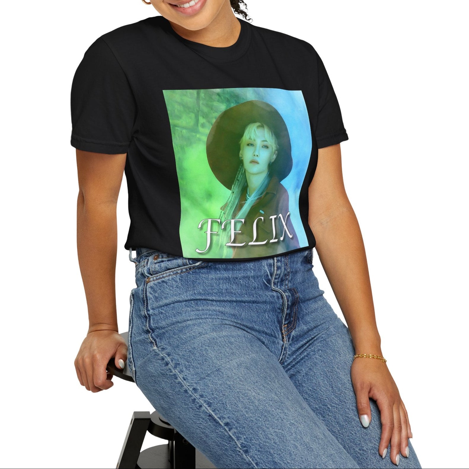 Model sitting on a stool and showcasing the unisex 100% ring-spun cotton T-Shirt in black, featuring a portrait of Stray Kids' Felix wearing a large hat. It is as if he was painted on canvas with the effect of a mist, blending soft light blue and light green tones. From ModeK Vetements.