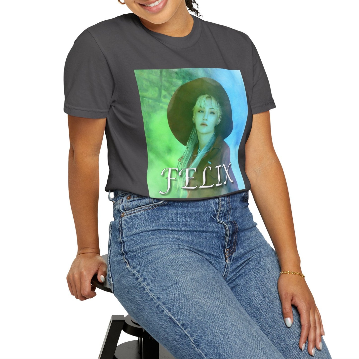 Model sitting on a stool and showcasing the unisex 100% ring-spun cotton T-Shirt in graphite, featuring a portrait of Stray Kids' Felix wearing a large hat. It is as if he was painted on canvas with the effect of a mist, blending soft light blue and light green tones. From ModeK Vetements.