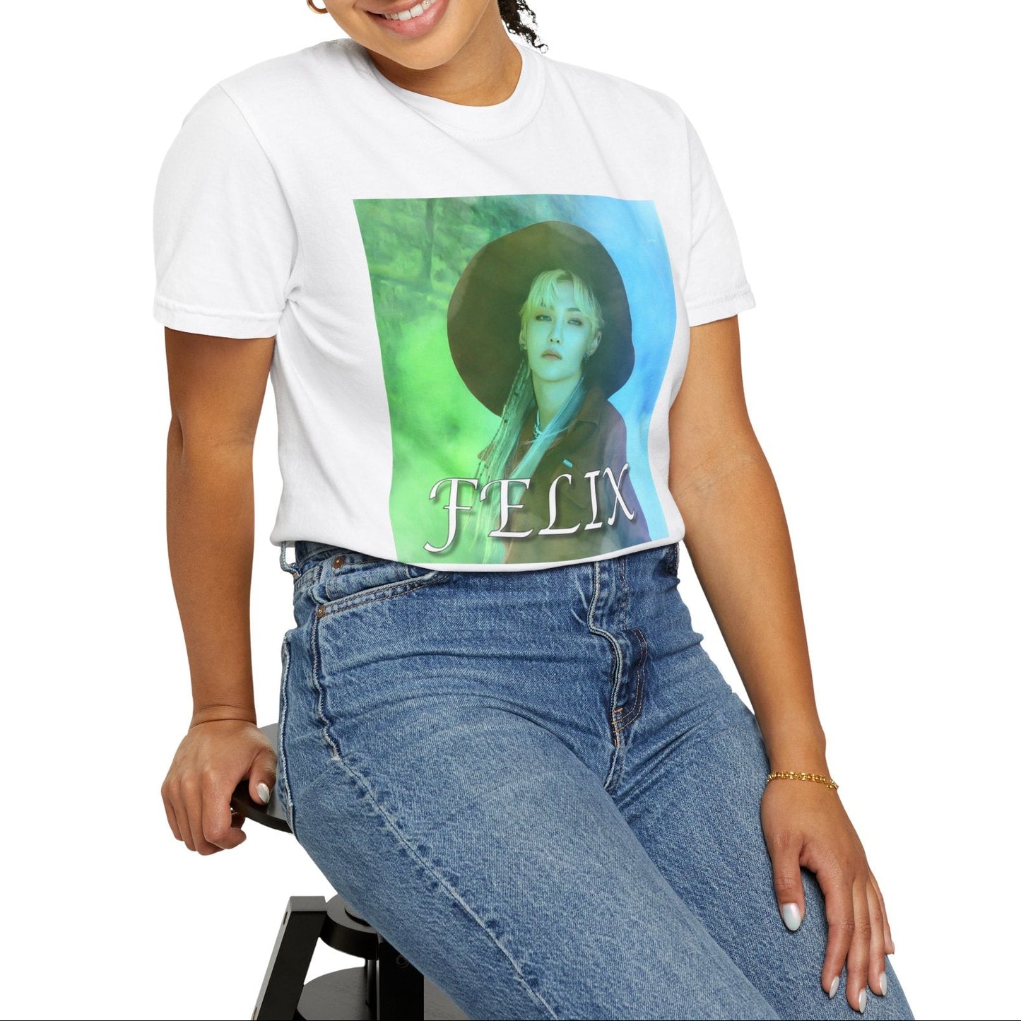 Model sitting on a stool and showcasing the unisex 100% ring-spun cotton T-Shirt in white, featuring a portrait of Stray Kids' Felix wearing a large hat. It is as if he was painted on canvas with the effect of a mist, blending soft light blue and light green tones. From ModeK Vetements.